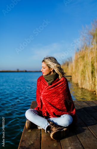 Young woman sitting on a wooden dock by the lake wrap around in a red blanket.