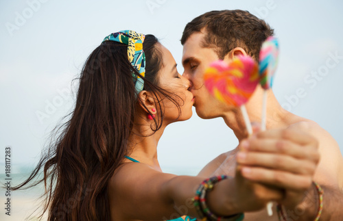 Summer lifestyle close up portrait of happy kissing couple in love. Holding in their hands lollipops in shape of heart. On honeymoon. Interracial couple, Asian woman, Caucasian man. Bright colors 