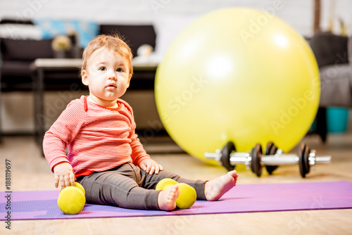 Portrait of a baby boy sitting on the mat with dumbbells and fitness ball at home