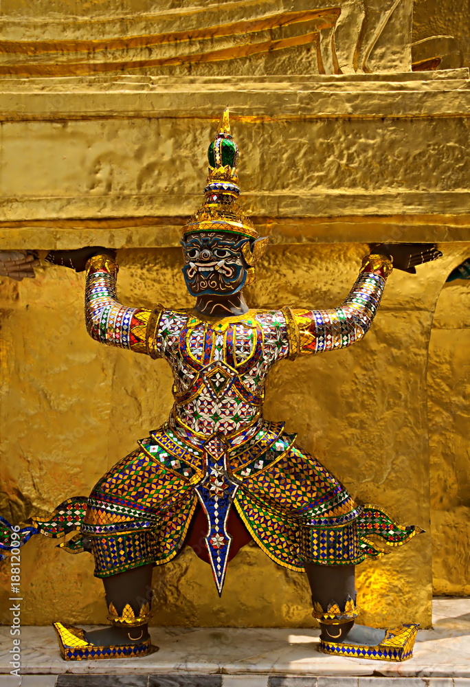 Close up of a colorful demon statue surrounding the base of a golden chedi at Wat Phra Kaew, Bangkok, Thailand. It appears as if he is holding up the chedi.