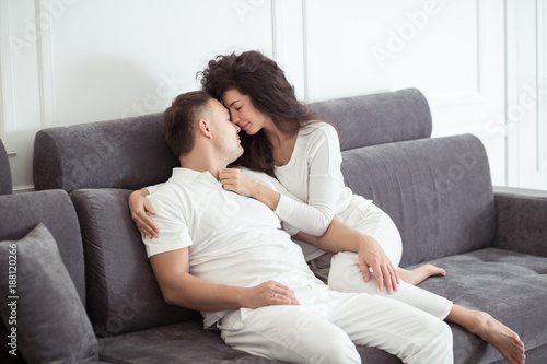 Portrait of young couple sitting together on grey sofa in modern white apartment or domestic room. Happy lovely family embracing and hugging together close-up © uv_group