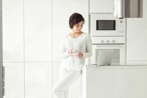 Young real beautiful woman planning expenses and paying bills on her white modern kitchen with laptop.