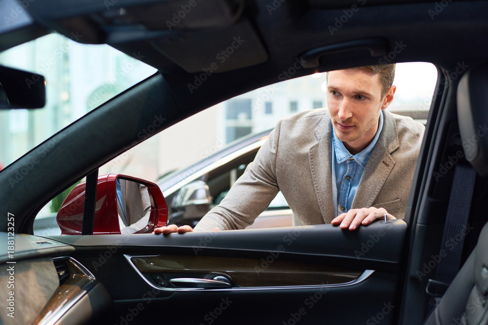 Portrait of handsome young man choosing new car in showroom looking inside open window at car interior Portrait of handsome young man choosing new car in showroom looking inside open window at car interior