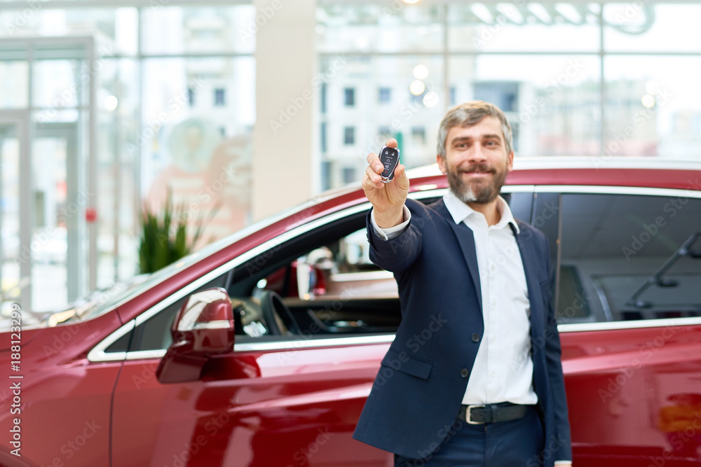Portrait of handsome mature man smiling happily at camera standing by brand new luxury car in showroom and presenting car keys, copy space