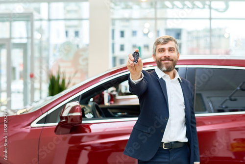 Portrait of handsome mature man smiling happily at camera standing by brand new luxury car in showroom and presenting car keys, copy space