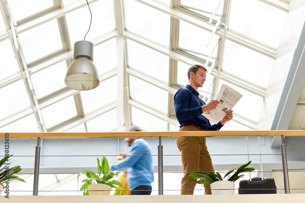 Low angle portrait of young businessman wearing casual clothes reading newspaper standing at balcony in modern office building under glass roof Low angle portrait of young businessman wearing casual clothes reading newspaper standing at balcony in modern office building under glass roof