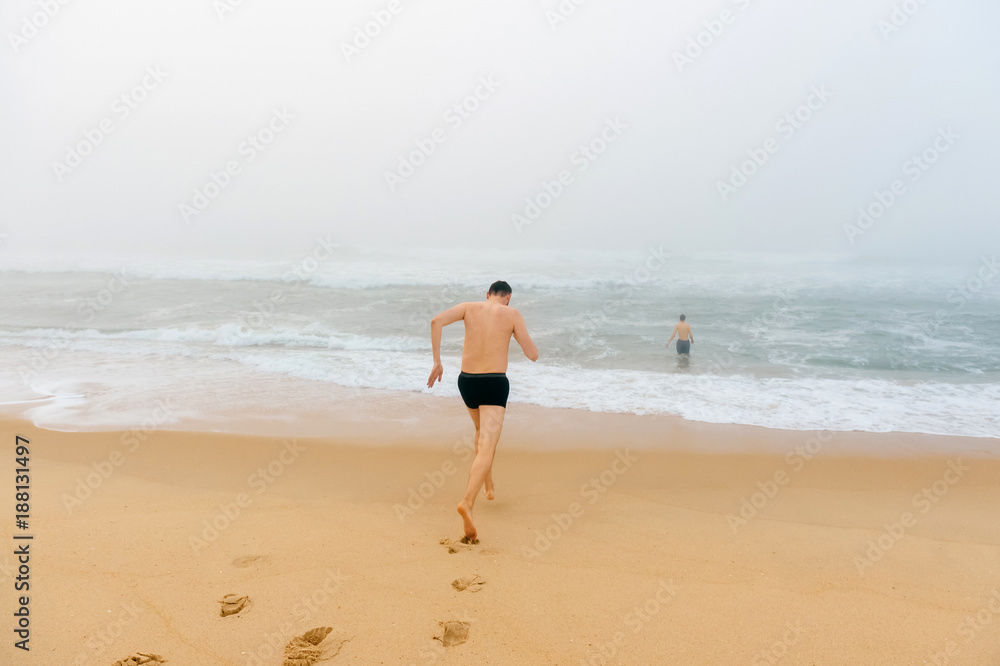 Topless man running from sandy beach in misty stormy ocean. Adult male enjoying swimming like child. Happy unrecognizable person. Water procedures. Healthcare and recreation. Crazy guy on vacation. Topless man running from sandy beach in misty stormy ocean. Adult male enjoying swimming like child. Happy unrecognizable person. Water procedures. Healthcare and recreation. Crazy guy on vacation.