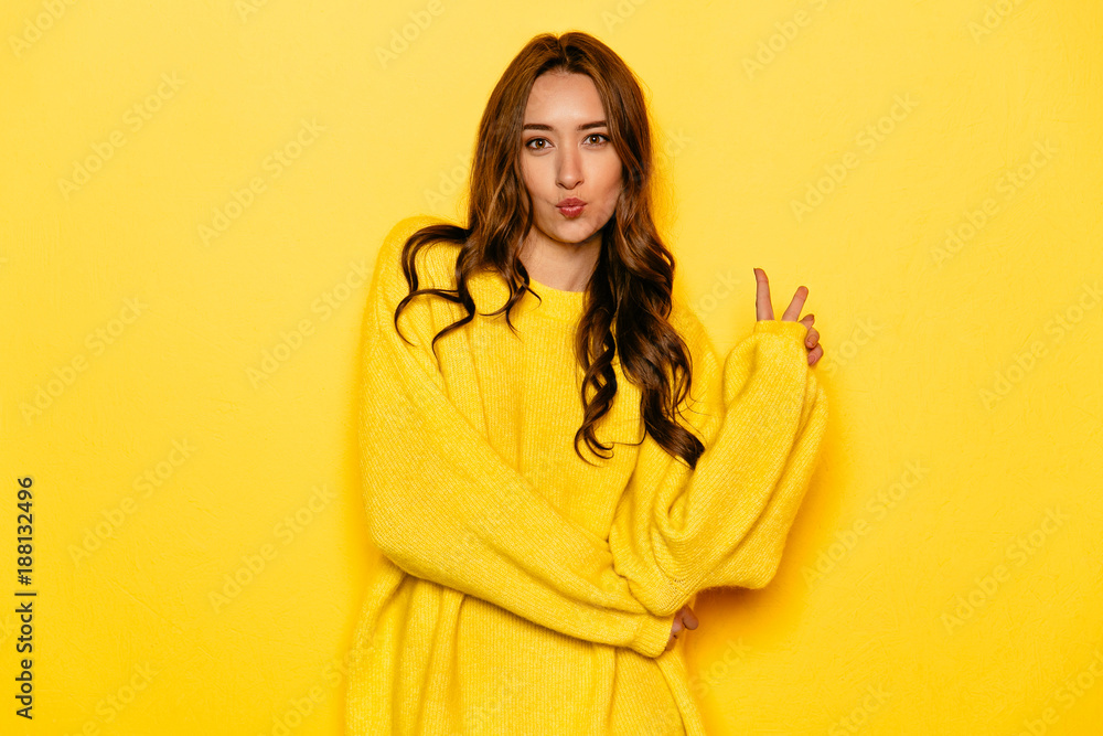 Charming young woman with long curly hair showing peace gesture, giving a kiss, dressed in yellow sweater.Yellow background. Charming young woman with long curly hair showing peace gesture, giving a kiss, dressed in yellow sweater.Yellow background.