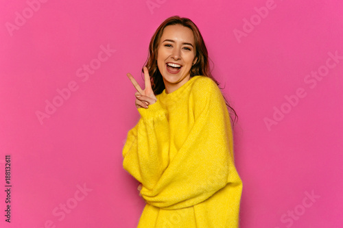 Gorgeous young woman showing peace gesture, dressed in yellow sweater. Over pink background. Concept of Valentine's day. © Maksym Azovtsev