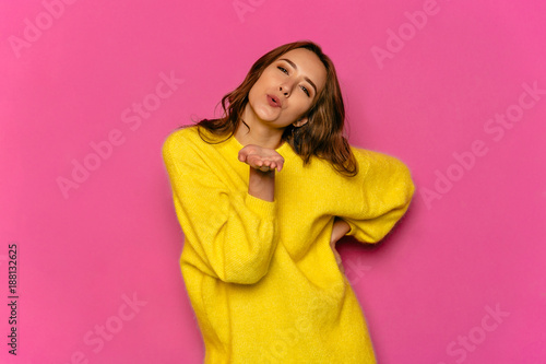 Concept of Valentine's day. Young pretty woman, dressed in yellow soft sweater, giving air kiss, looking at camera. Isolated on pink background. © Maksym Azovtsev