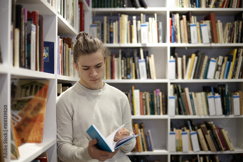 People, learning, knowledge, education and school concept. Serious young guy wearing sweater standing in bookstore, reading extract from textbook in his hands, leaning on white shelves full of books People, learning, knowledge, education and school concept. Serious young guy wearing sweater standing in bookstore, reading extract from textbook in his hands, leaning on white shelves full of books