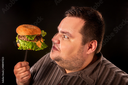 Diet failure of fat man eating fast food. Overweight person eating huge hamburger on fork. Junk meal leads to obesity. Man studies quality of product before eating.