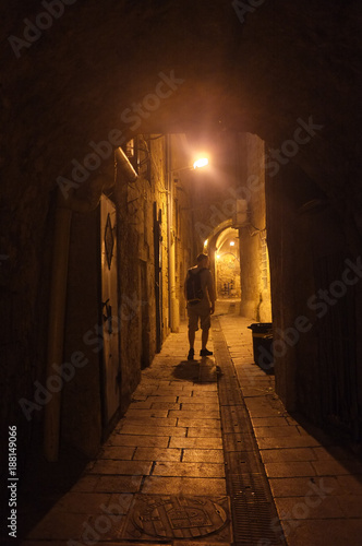 Illuminated cobbled street with light reflections on cobblestones in old historical city by night. Dark blurred silhouette of person goes in search of adventure in Old Jaffa in Tel-Aviv, Israel