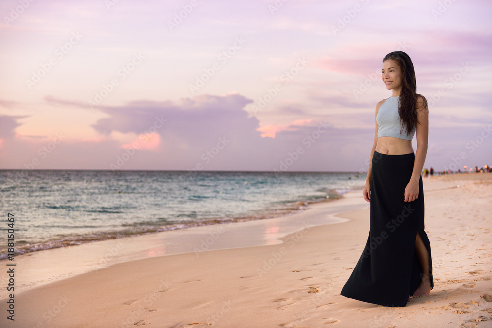 Asian model woman walking on beach at sunset relaxing on luxury Caribbean travel holiday wearing fashion maxi skirt outfit for night out on summer vacation. Chinese girl enjoying view of ocean.