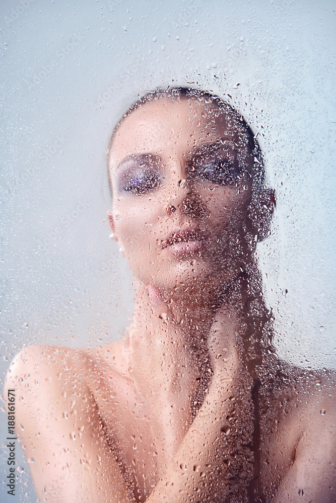 Brunette woman with makeup smoky eyes behind glass with water drops close-up