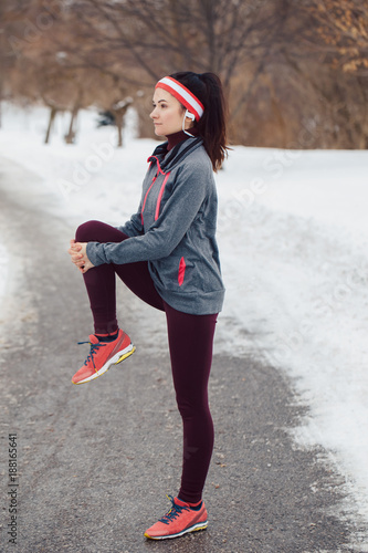 Portrait of Caucasian young woman stretching and warming up for jogging outside in winter park. Girl exercising outdoors wearing sportswear, headband and headphones.