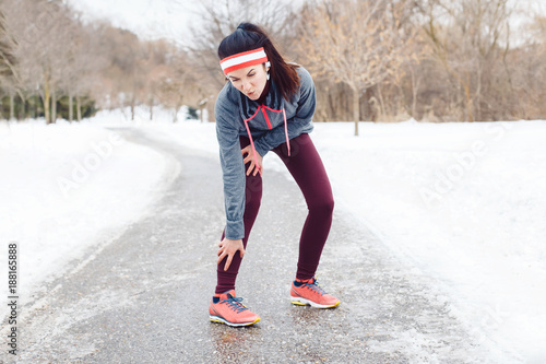Portrait of Caucasian young woman feeling pain in legs after jogging outside in winter park. Girl having injury when running excercising outdoors in cold weather conditions