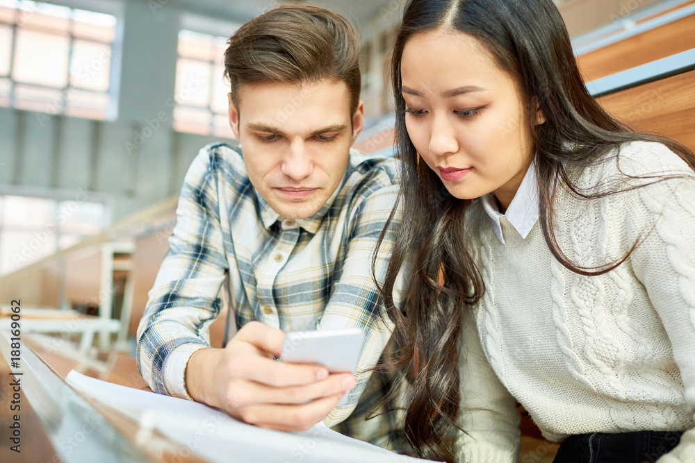 Portrait of two students, young man and Asian woman, using smartphone during break, sitting at desk in lecture hall of modern college, copy space Portrait of two students, young man and Asian woman, using smartphone during break, sitting at desk in lecture hall of modern college, copy space