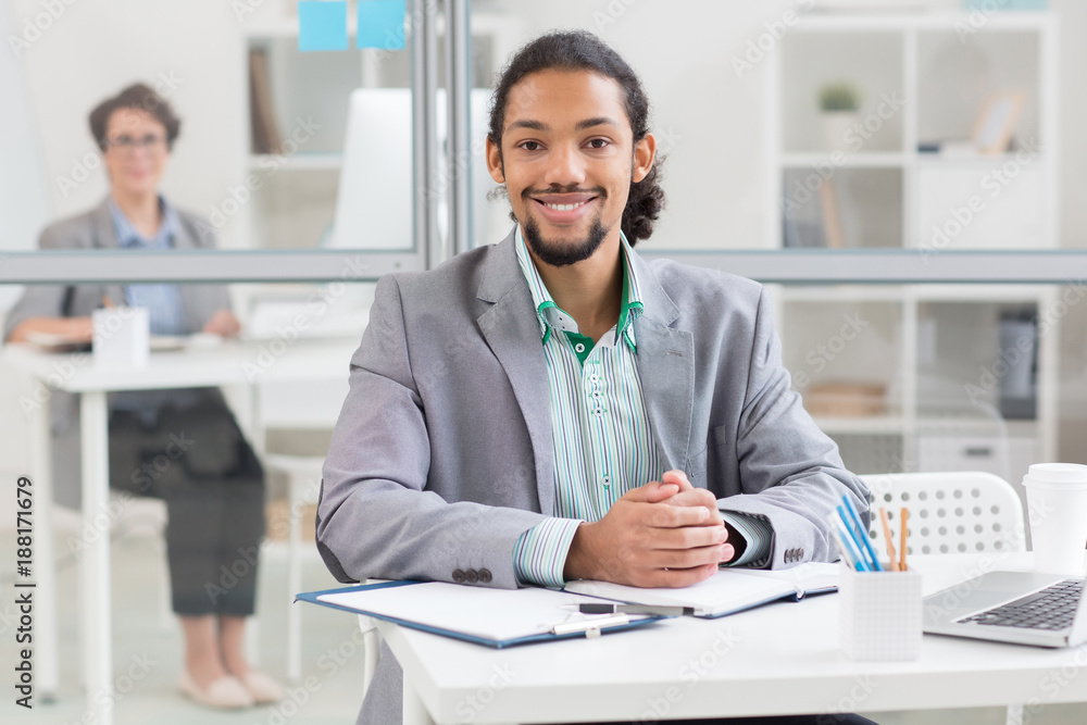 Successful African-american manager sitting by workplace in office and looking at camera with colleague on background Successful African-american manager sitting by workplace in office and looking at camera with colleague on background