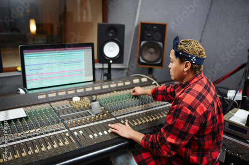 Young woman in shirt and headband sitting by stereo equipment in record studio and working