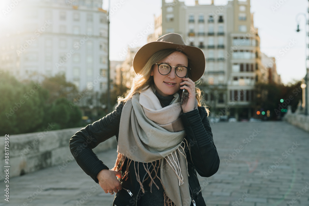 Sunny autumn day, backlight. Young attractive woman travels in hat and eyeglasses stands on city street, talking on cell phone, smiling. Hipster girl walks. Vacation, adventure, trip, traveling.