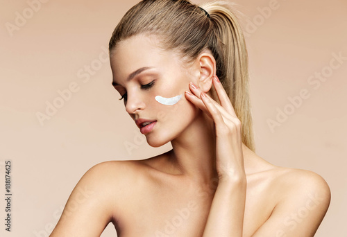 Gorgeous blonde girl applying moisturizing cream on her face. Photo of young girl with flawless skin on beige background. Skin care and beauty photo