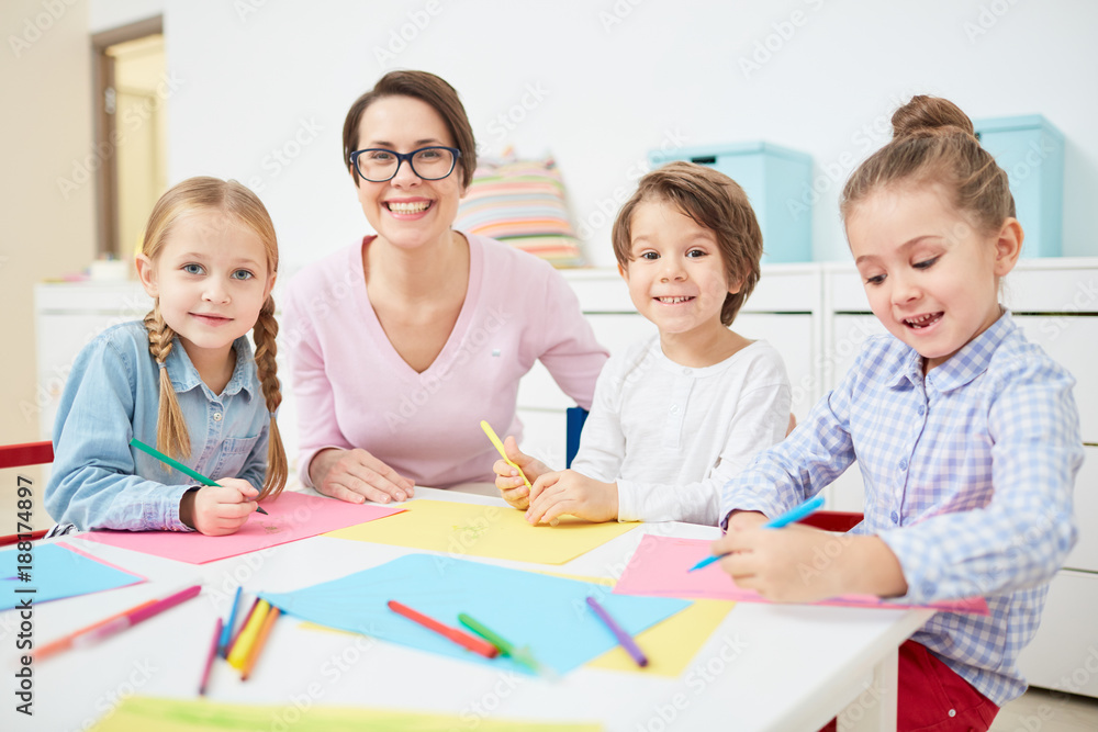 Happy young teacher and small group of adorable kids drawing with crayons in kindergarten