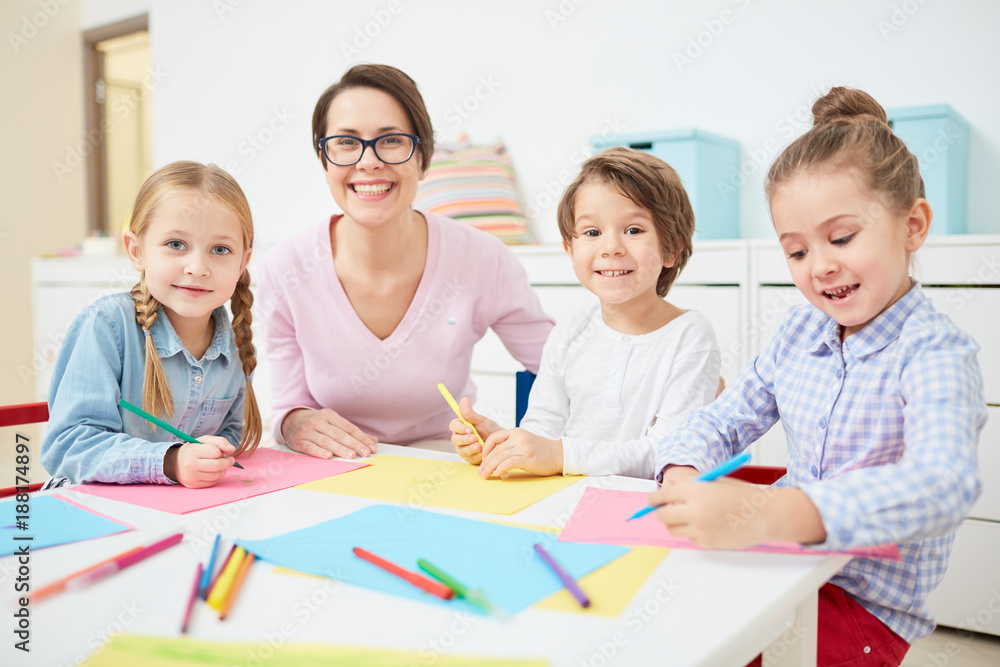Happy young teacher and small group of adorable kids drawing with crayons in kindergarten Happy young teacher and small group of adorable kids drawing with crayons in kindergarten