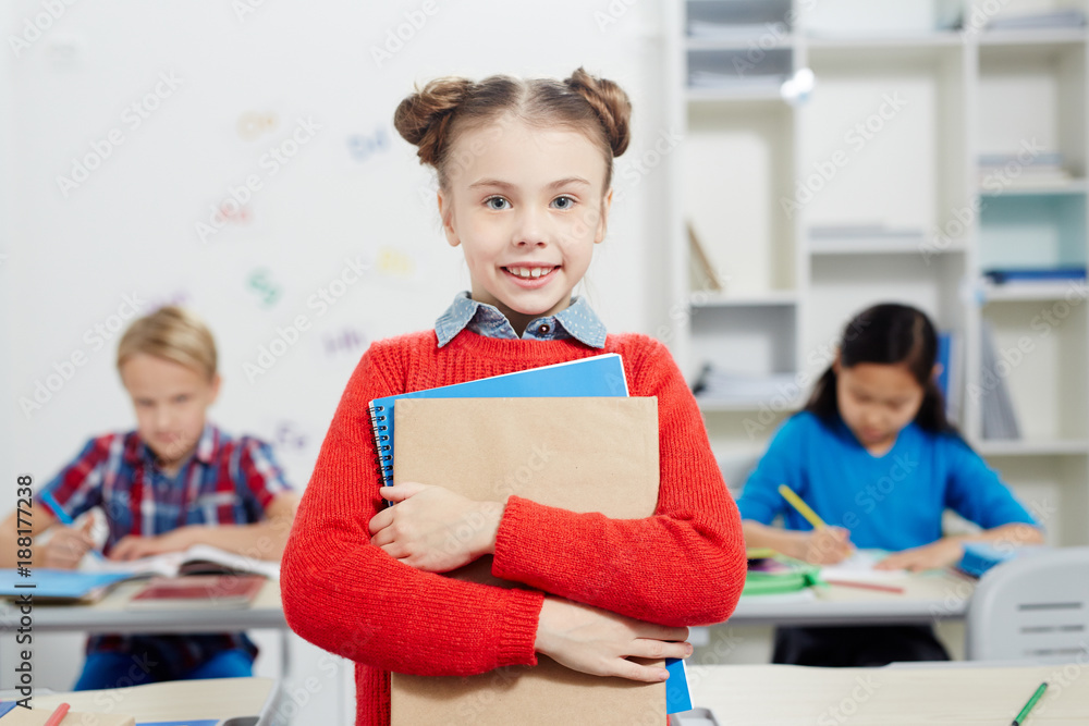 Pretty schoolgirl with books looking at camera with two classmates writing test on background Pretty schoolgirl with books looking at camera with two classmates writing test on background