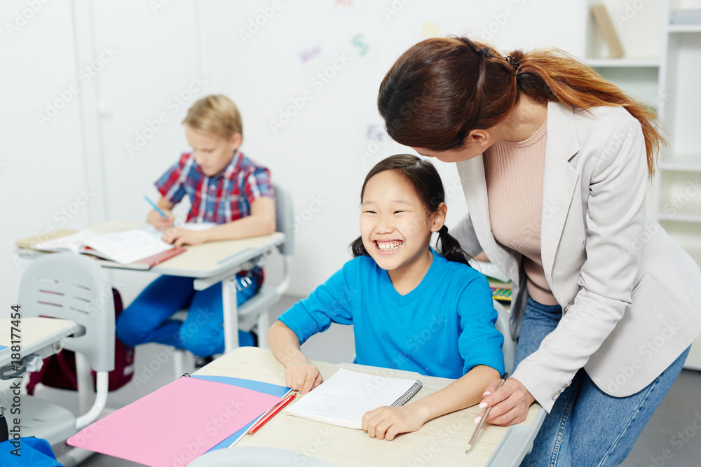 Laughing girl by desk looking at blackboard while listening to her teacher near by Laughing girl by desk looking at blackboard while listening to her teacher near by