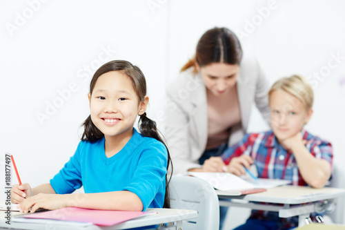 Youthful girl with crayon looking at camera with teacher and schoolboy talking on background