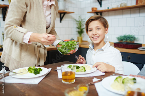 Happy boy looking at camera by dinner while his grandmother putting him vegetable salad on plate