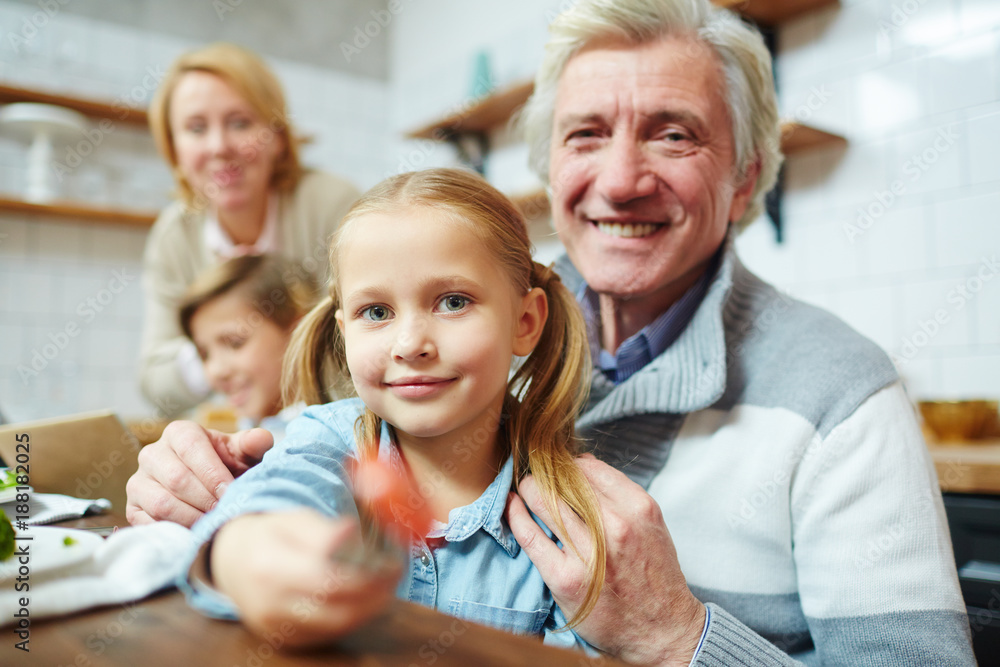 Cute little girl offering you fresh tomato slice on fork with her grandfather near by Cute little girl offering you fresh tomato slice on fork with her grandfather near by