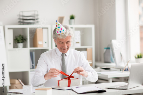 Happy senior businessman in birthday-cap untying ribbon on top of gift-box in office
