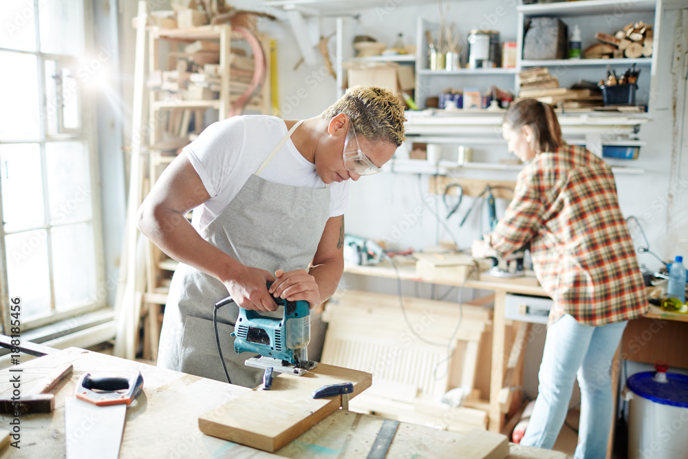 Fototapeta premium Young carpenter with electric fretsaw sawing wooden board in workshop