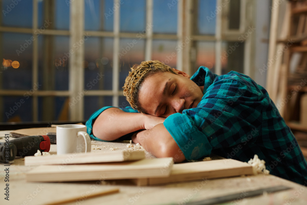 Exhausted carpenter sleeping by workplace with mug and wooden materials near by Exhausted carpenter sleeping by workplace with mug and wooden materials near by