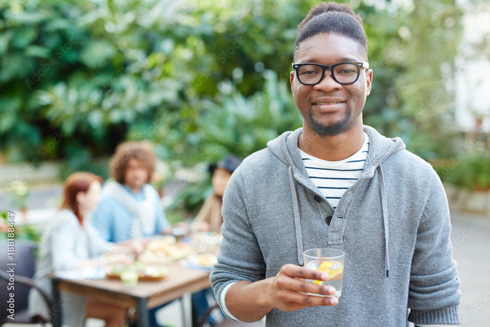 African guy with refreshing drink looking at camera with friends having dinner on background African guy with refreshing drink looking at camera with friends having dinner on background