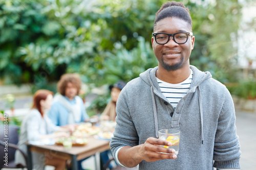 African guy with refreshing drink looking at camera with friends having dinner on background