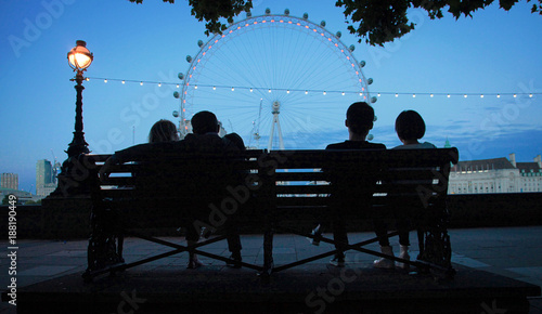 Romantic and atmospheric panoramic image of London at night, featuring four silhouetted people on a bench looking out across the South Bank, London