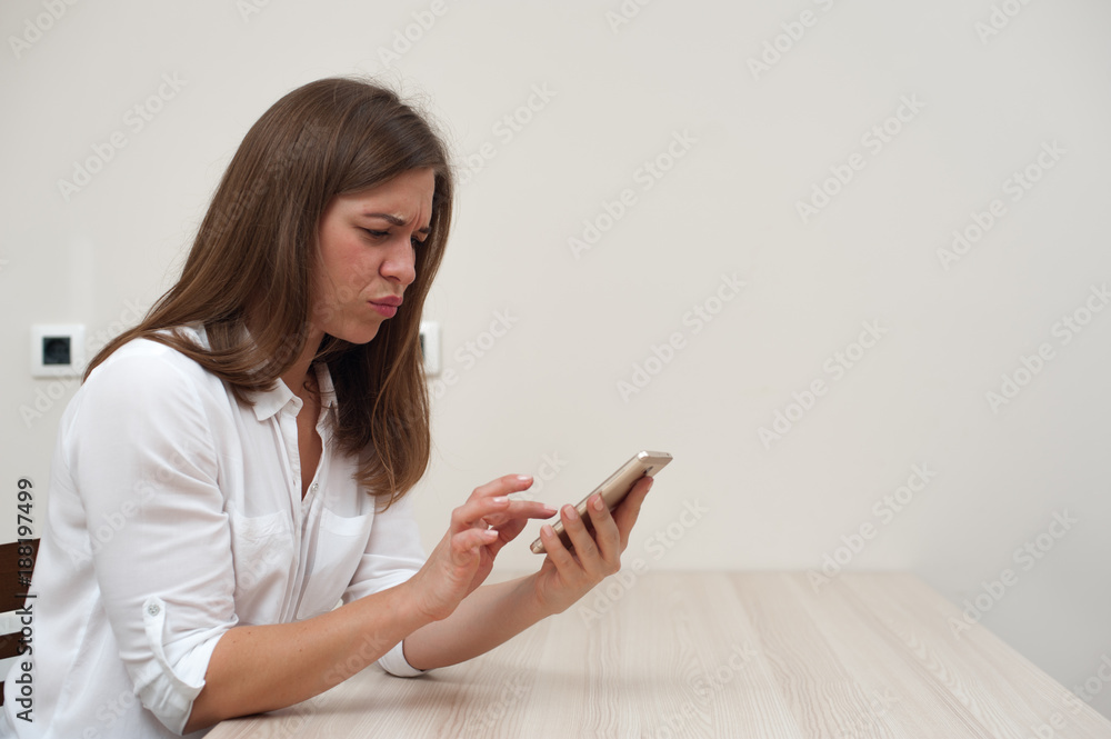 Displeased angry young woman wearing white shirt using her smartphone and looking into the screen. Copyspace. Displeased angry young woman wearing white shirt using her smartphone and looking into the screen. Copyspace.