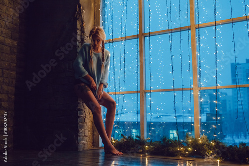 Sexy blond girl in a sweater stands near a window.