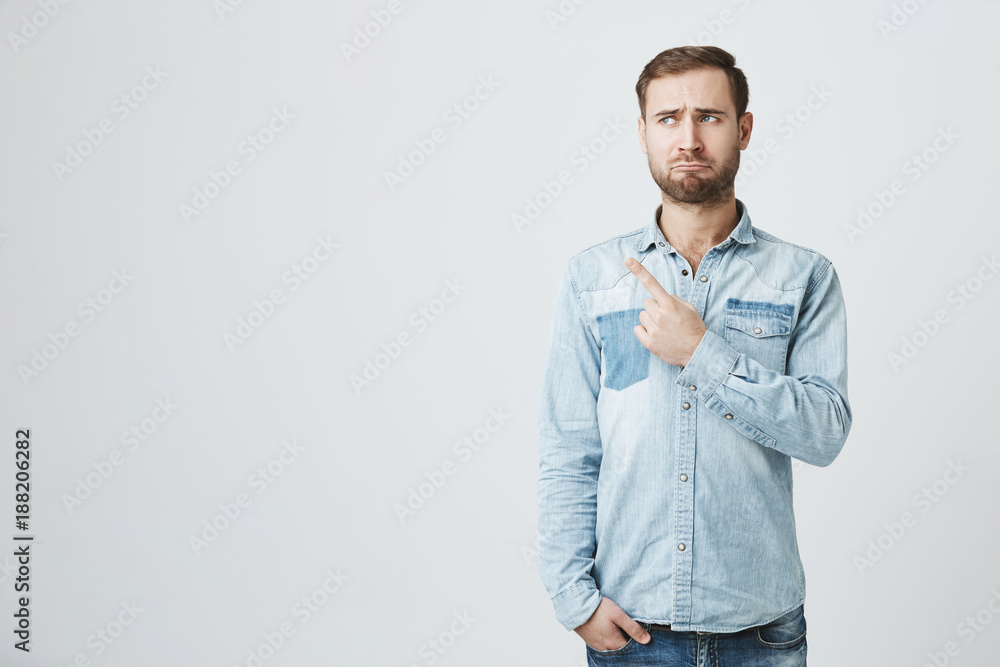 Upset european bearded man wearing denim shirt frowning face looking sadly at camera, pointing finger at copy space on studio background. Caucasian handsome man indicating something on wall Upset european bearded man wearing denim shirt frowning face looking sadly at camera, pointing finger at copy space on studio background. Caucasian handsome man indicating something on wall