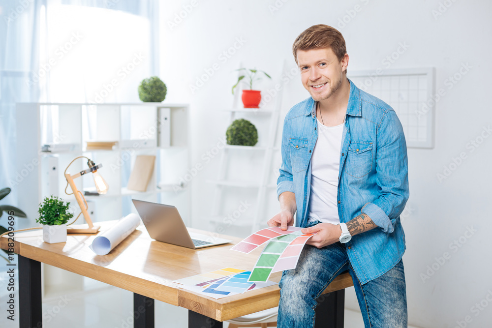 Color palettes. Cheerful handsome positive designer sitting on the table and smiling while holding bright color palettes