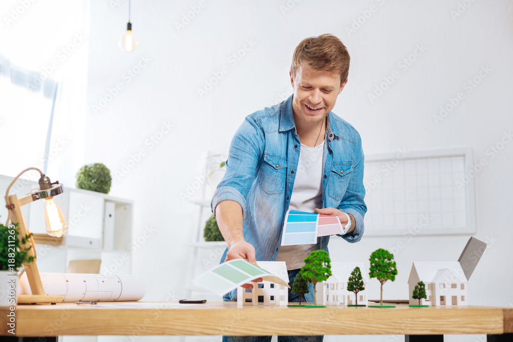 Suitable colors. Attentive responsible young engineer having a productive day at work while standing near his table and choosing a suitable color for new houses Suitable colors. Attentive responsible young engineer having a productive day at work while standing near his table and choosing a suitable color for new houses