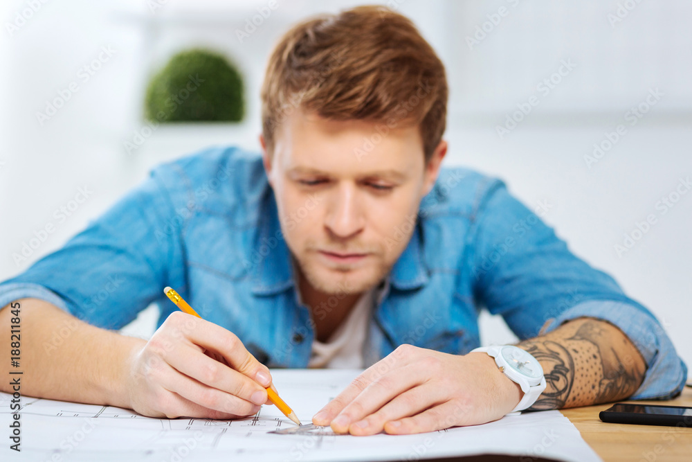 Very careful. Calm attentive smart engineer sitting at the table in his comfortable office and looking careful while drawing a straight line on a scheme Very careful. Calm attentive smart engineer sitting at the table in his comfortable office and looking careful while drawing a straight line on a scheme