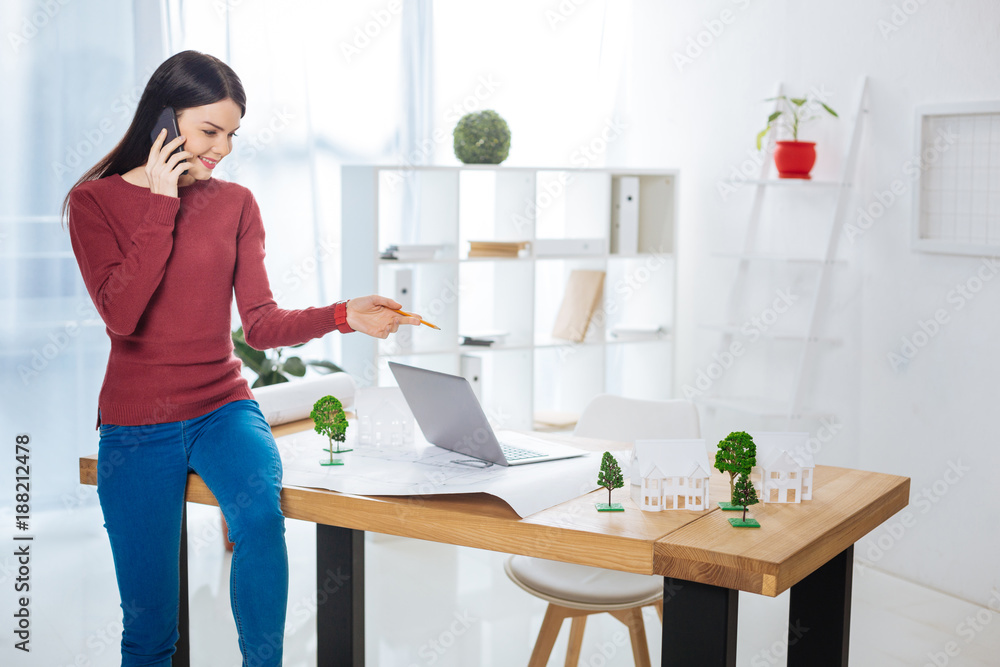 Friendly conversation. Cheerful young skilled engineer sitting on the table in a light comfortable office and feeling happy while talking on the phone Friendly conversation. Cheerful young skilled engineer sitting on the table in a light comfortable office and feeling happy while talking on the phone