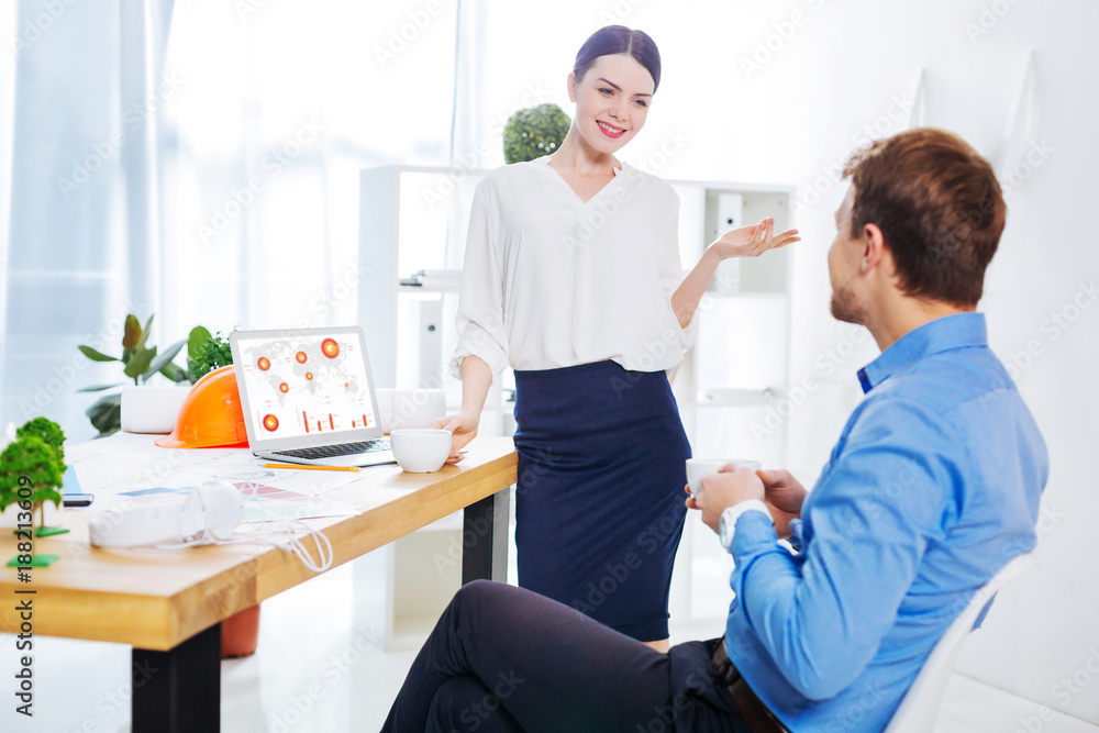Obraz premium Elegant colleague. Young pretty elegant woman looking friendly while standing in front of her smart colleague and looking happy while having a pleasant talk with him
