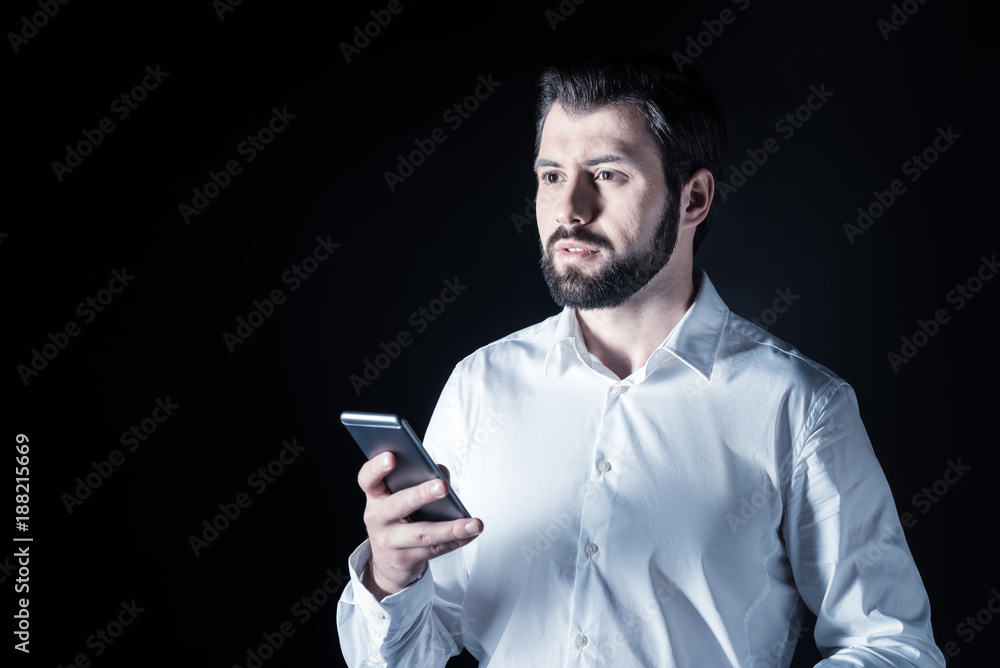 Modern gadget. Serious nice handsome man holding his smartphone and using it while standing against black background Modern gadget. Serious nice handsome man holding his smartphone and using it while standing against black background