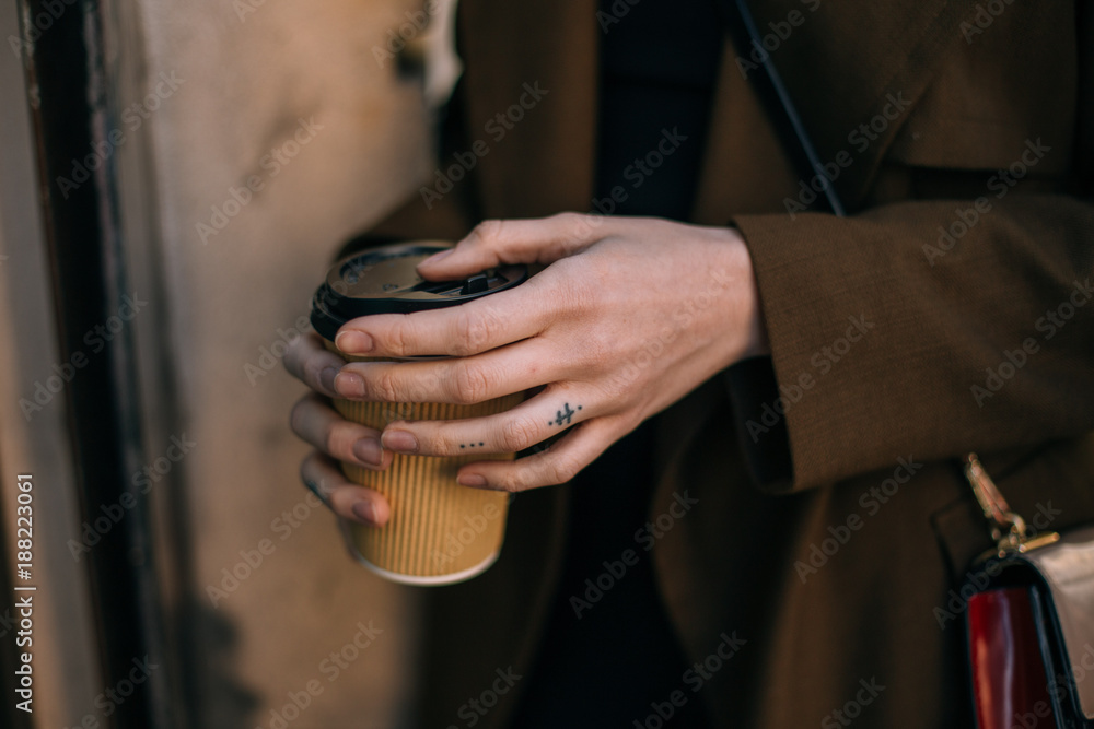 Fashionable woman in coat, stands outdoors on street, holds in hand with tattooed fingers, take away cup with coffee to go. Vintage look and feel nostalgia photo, tender and sensual morning routine