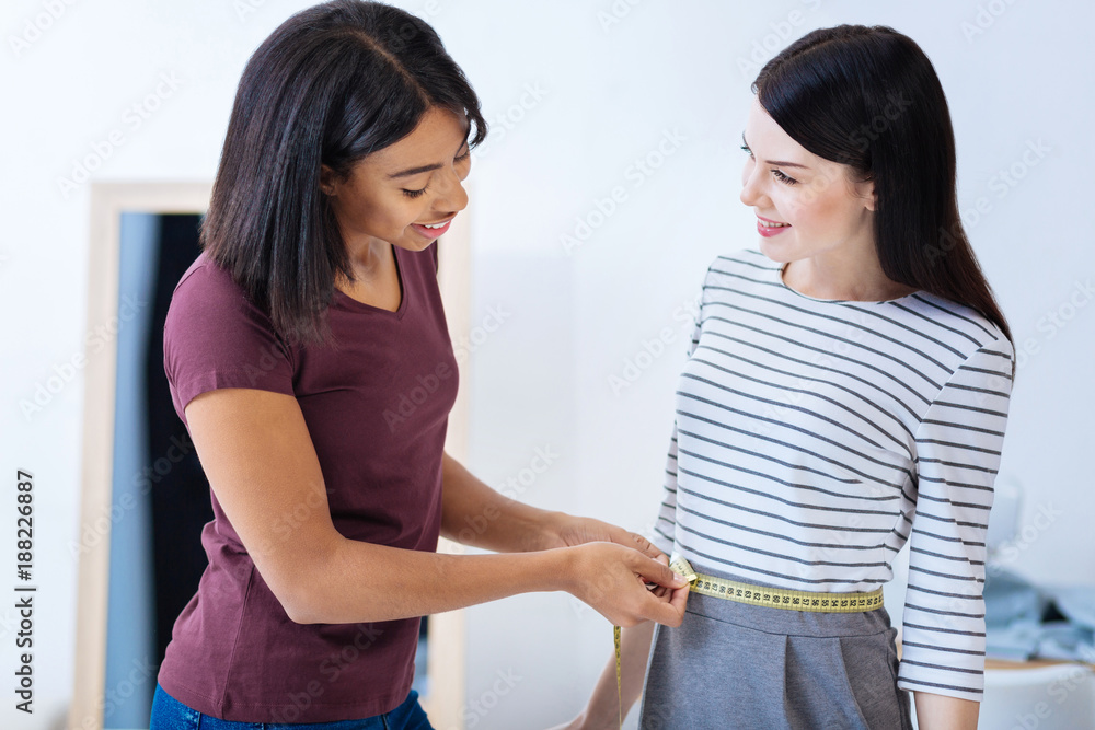 Slim person. Calm professional attentive tailor carefully measuring the waist line of her curious client while being at work in her comfortable atelier Slim person. Calm professional attentive tailor carefully measuring the waist line of her curious client while being at work in her comfortable atelier
